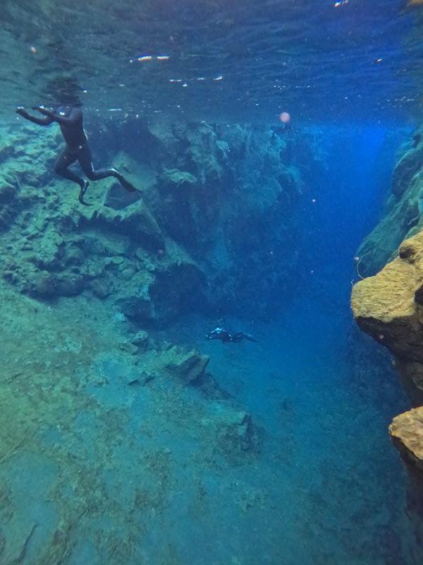 A freediver descending along the rocky wall of the Silfra fissure in blue glacial water