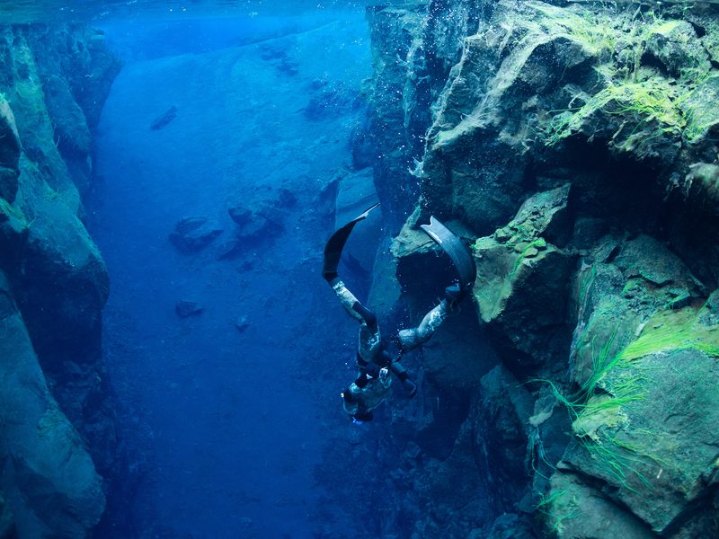 A freediver descending headfirst along a mossy rock wall in Silfra