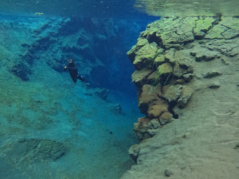 A freediver gliding past a moss-covered rock ledge in Silfra